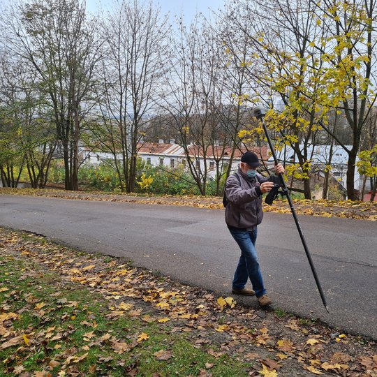 Nuotraukoje žilas žmogus, kuris yra su juodos spalvos kepure, pilkos spalvos striuke, mėlynais džinsais ir smėlio spalvos batais. Jis taip pat užsidėjęs šviesiai mėlynos spalvos veido kaukę. Vyras rankose laiko juodos spalvos matavimo prietaisą. Už jo deš