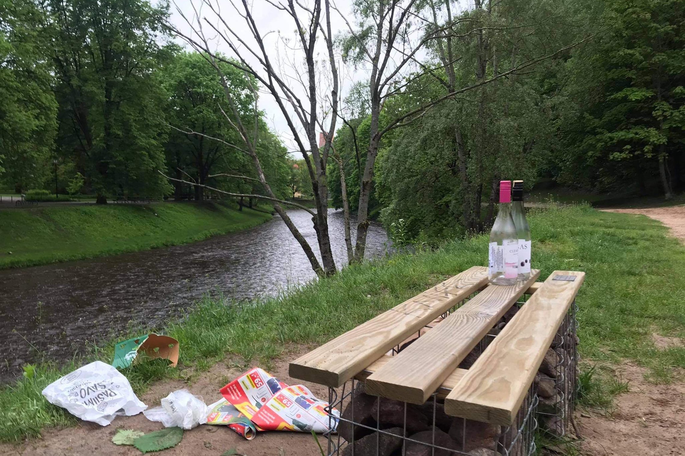 Vilnios krantas. Stovi medinis suoliukas. Aplink išverstos šiukšlės, padėtas butelis ant suolo. Yra vasara, žali Vilnios pakrantės medžiai.