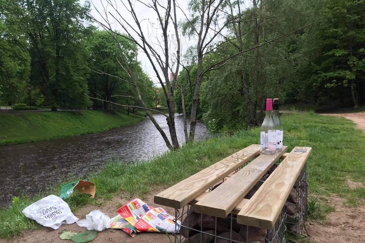 Vilnios krantas. Stovi medinis suoliukas. Aplink išverstos šiukšlės, padėtas butelis ant suolo. Yra vasara, žali Vilnios pakrantės medžiai.