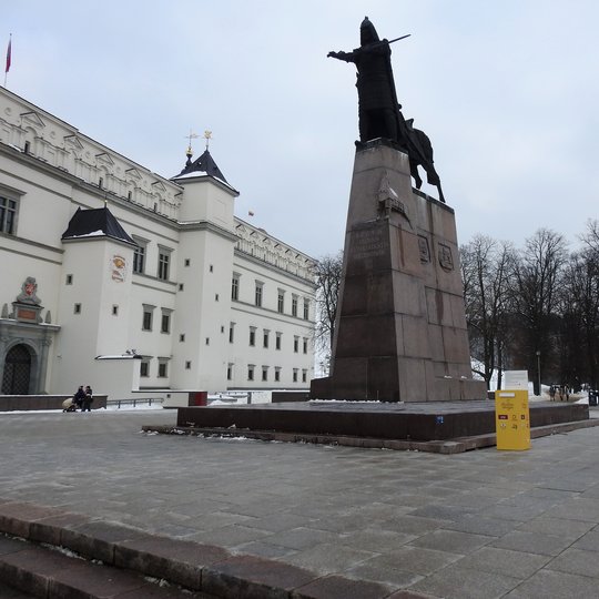 Kunigaikščio Gedimino paminklas, kurio papėdėje stovi geltona laiškų dėžutė. Už paminklo stūkso balti Valdovų rūmai ir Pilių parko medžiai.