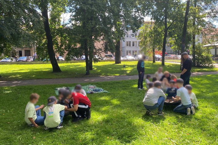 Vaikai su vadovu lauke, žalioje pievelėje, susėdę grupelėmis dalyvauja edukacijoje prie ugnies indo