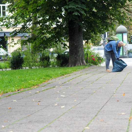 Du žmonės tvarko šaligatvį parke – vienas šluoja, kitas laiko šiukšlių maišą.
