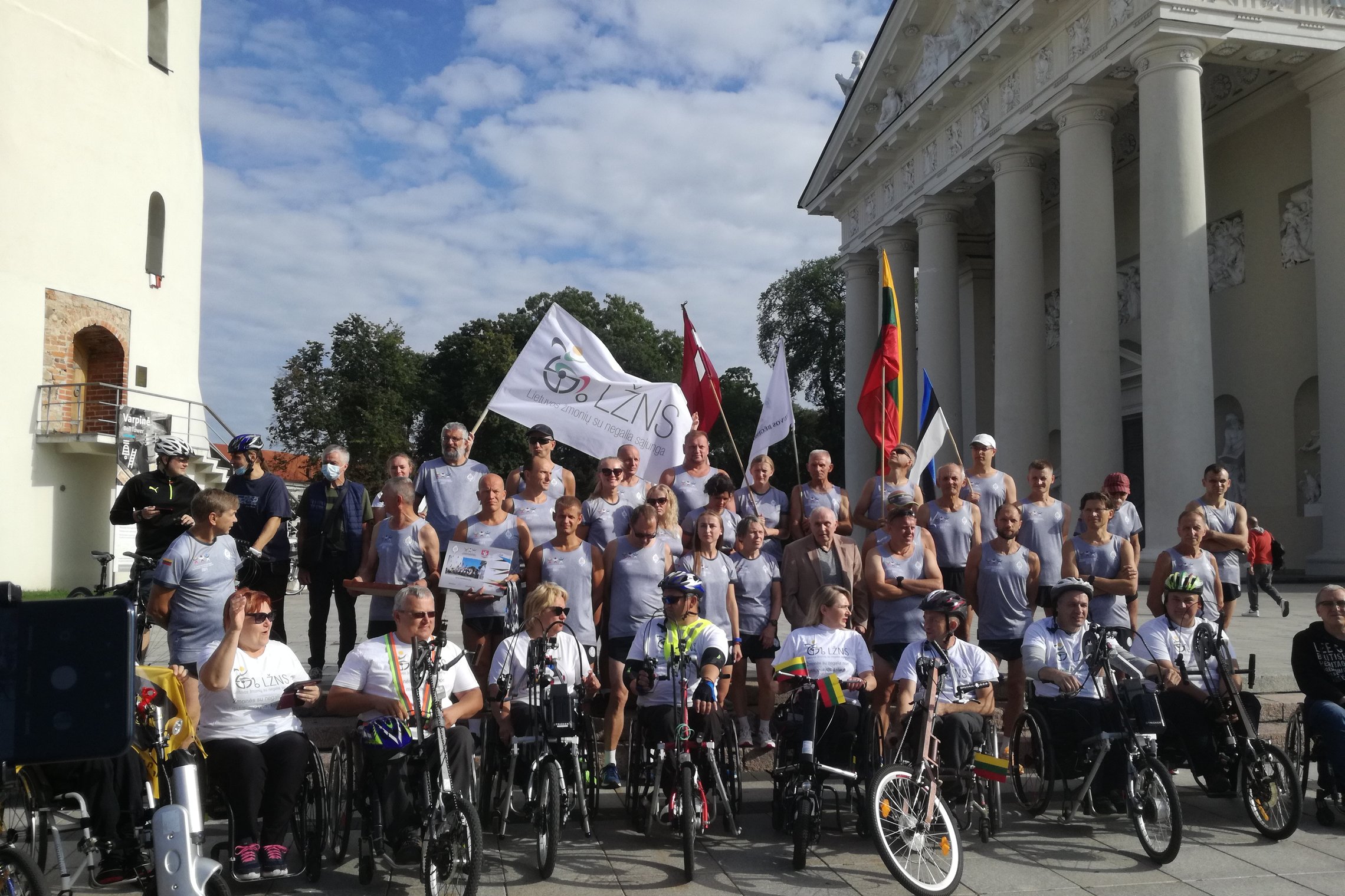 Tradiciško jubiliejinio estafetinio bėgimo dalyviai. Jie sėdi žmonių su negalia vežimėliuose. Jie nuotraukoje pozuoja tarp Katedros varpinės ir Katedros. Daug vėliavų, vienoda bėgikų apranga, labai graži diena, daug saulės.