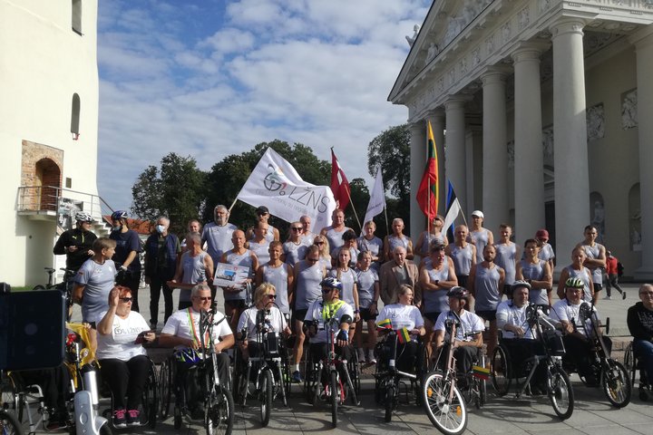 Tradiciško jubiliejinio estafetinio bėgimo dalyviai. Jie sėdi žmonių su negalia vežimėliuose. Jie nuotraukoje pozuoja tarp Katedros varpinės ir Katedros. Daug vėliavų, vienoda bėgikų apranga, labai graži diena, daug saulės.