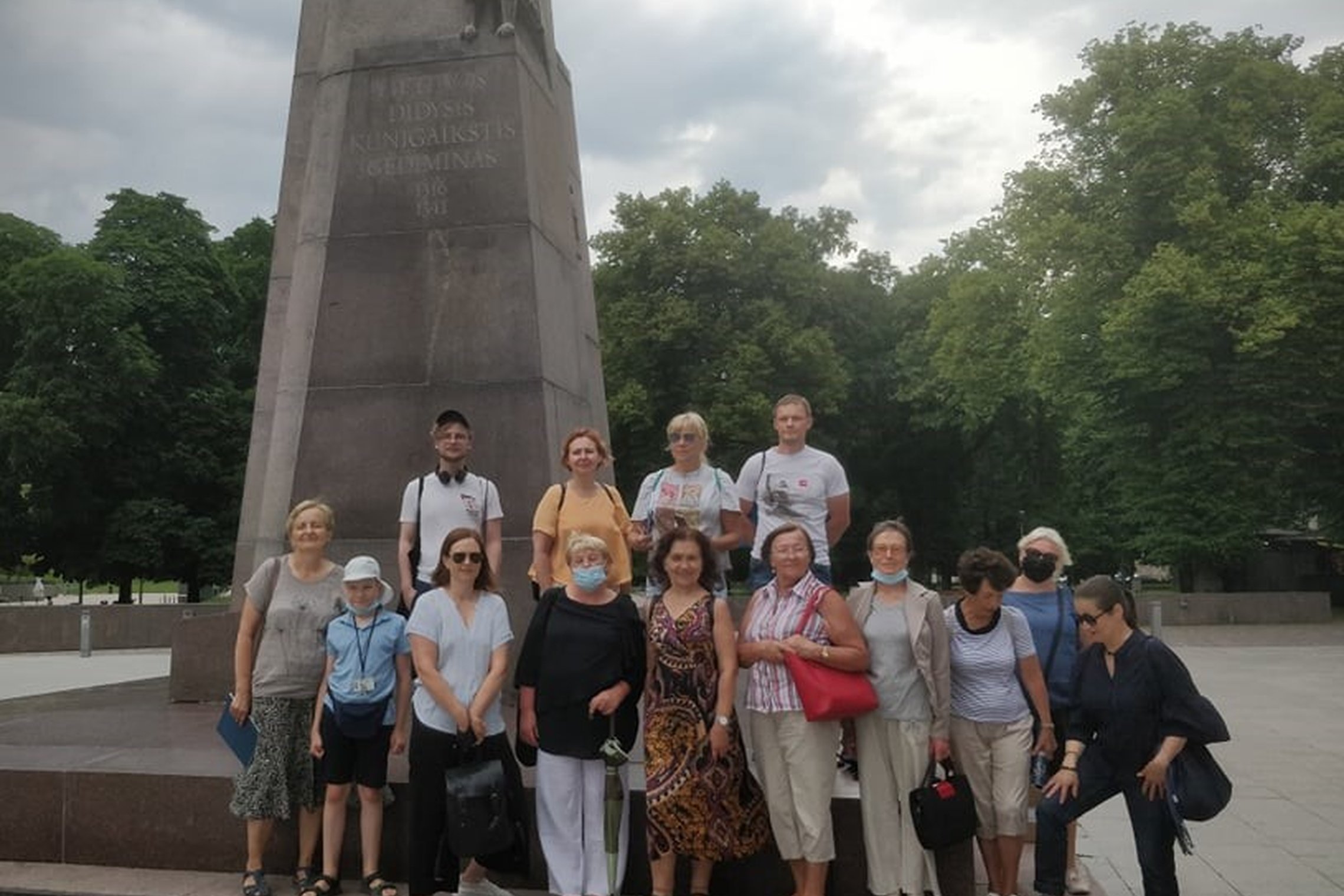 Didelė eksursijos grupė susirinkusi pozuoja Gedimino paminklo papėdėjė. Nors vasara, tačiau tamsi, debesuota diena.