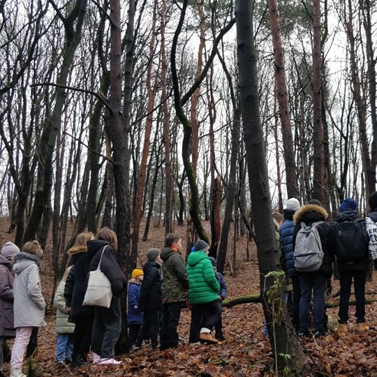 Nemažas būrelis vaikų tarp medžių. Vyksta ekskursija ir jie įdėmiai klausosi. Vaikai su stiukėmis, šiltai apsirengę, kai kurie turi kuprines.