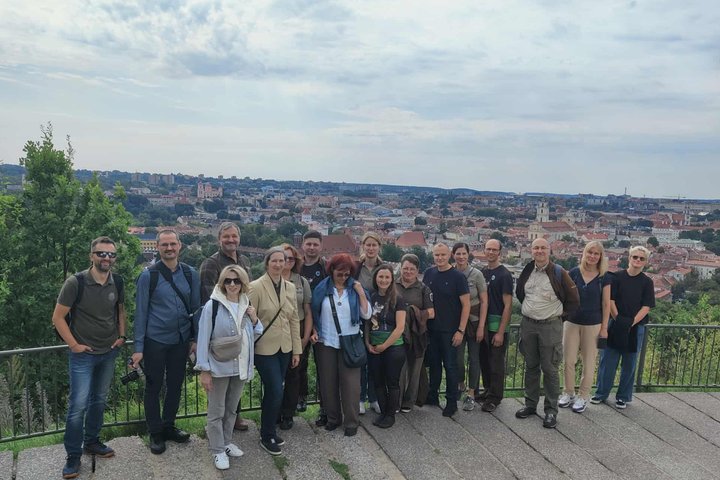 Grupė saugomų teritorijų darbuotojų stovi Vilniaus pilių rezervate ant kalno, o už jų nugarų atsiveria plati Vilniaus senamiesčio panorama su raudonų stogų jūra.