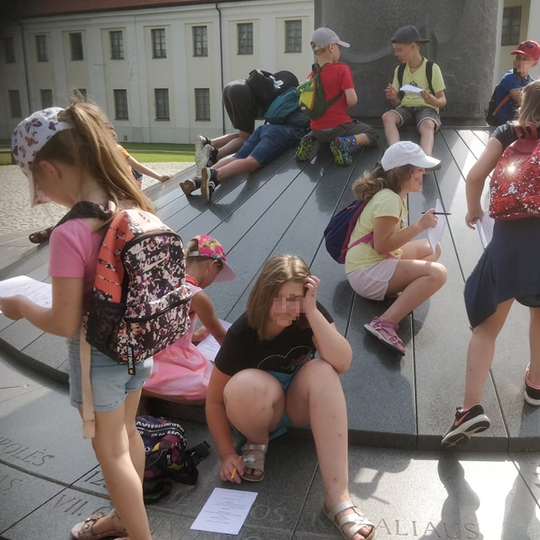 Vaikai sutupę aplink Mindaugo paminklą sprendžia žaidimo užduotis. Yra karšta vasaros diena. Vaikai apsirengę vasariškai.