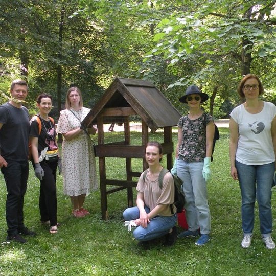 Žmonių grupė stovi ir pozuoja prie medinės lesyklos parke.