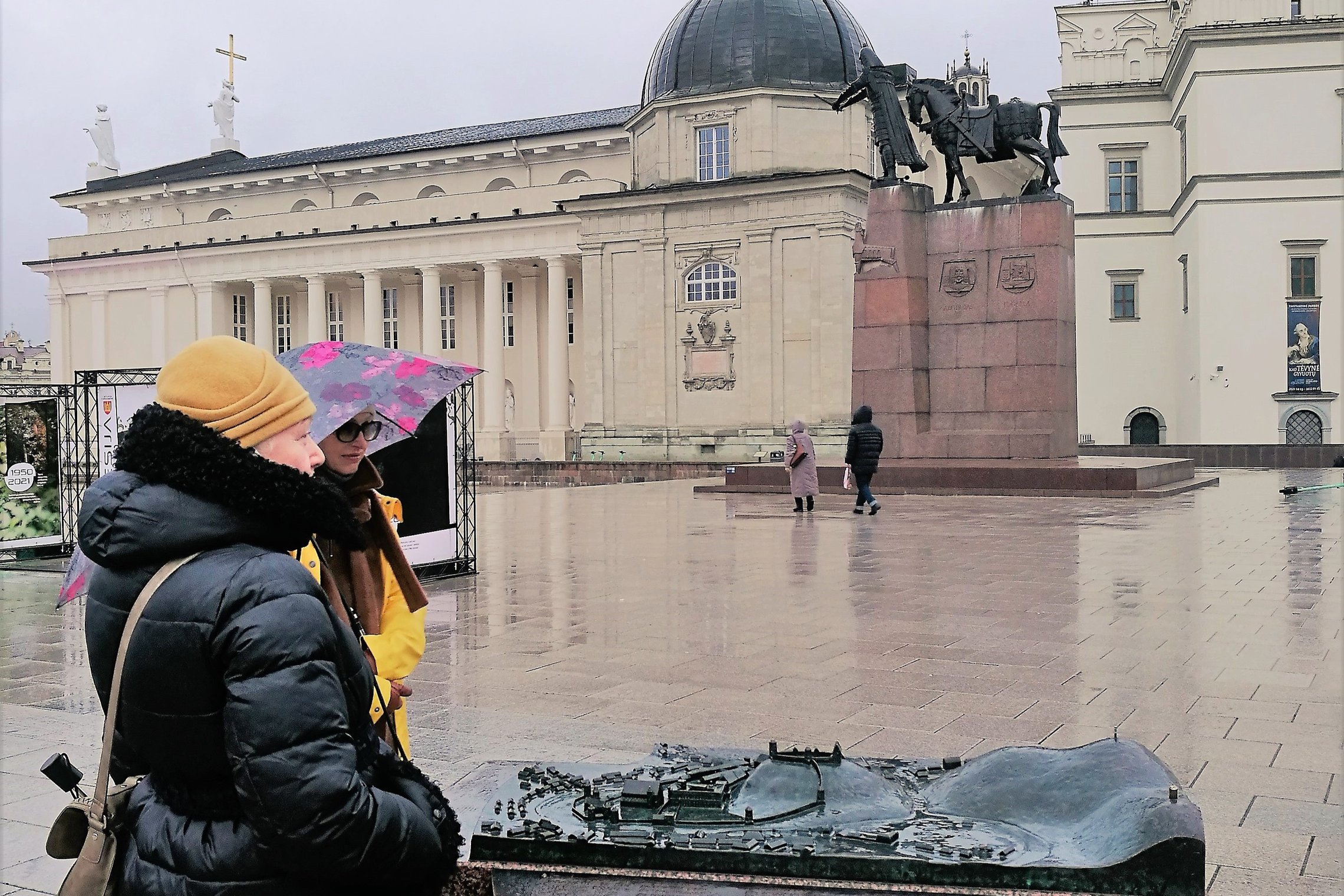 Vilniaus katedros aišktė ir joje esantis Vilniaus pilių bronzinis maketas. Dvi moterys tyrinėja makete esančius objektus. Dešinėje matosi Valdovų rūmai ir Vilniaus katedra su Šv. Kazimiero koplyčia. Lyja lietus, nes moterys su lietsargiu.