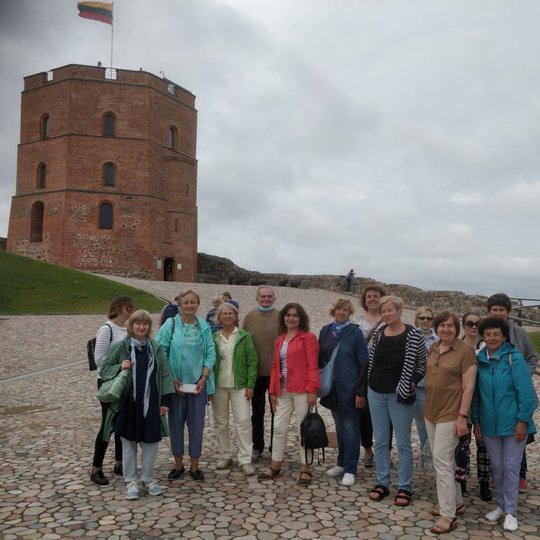 Nemaža ekskursijos grupė pozuoja nuotraukai Gedimino pilies bokšto papėdėje. Ekskursijos dalyviai stovi ant gražaus naujai įrengto akmeninio grindinio.