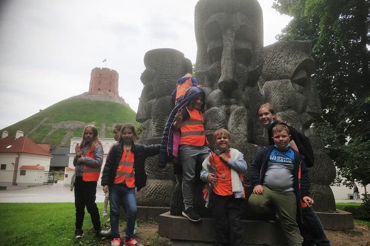 Ryškiomis, šviesą atspindinčiomis liemenėmis dėvintys vaikai žaidžia orientacinį žaidimą Vilniaus pilių rezervate. Antrame nuotraukos fone matosi Gedimino pilies kalnas ir bokštas.