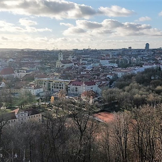 Nuostabi ir šilta rudens diena ir nuotraukoje daug vaiskaus dangaus su baltais debesimis. Vaizdas nuo Trijų kryžių kalno. Apačioje teniso kortai ir visas Vilniaus miestas. Nuotraukos pirmame plane - daug medžių. Jie jau numetę lapus.