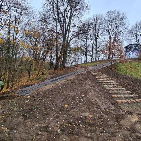 Dešinėje randasi pradėtų mūryti laiptų konstrukcija: laiptai lipinami iš akmenų ir cemento mišinio. Akmenys nemaži, vienoje pakopoje yra 5 – 6 akmenys. Laiptų konstrukcija lipdama į kalvelę pasiekia būsimą taką ir už jo tęsiasi toliau. Dešinėje apačioje l