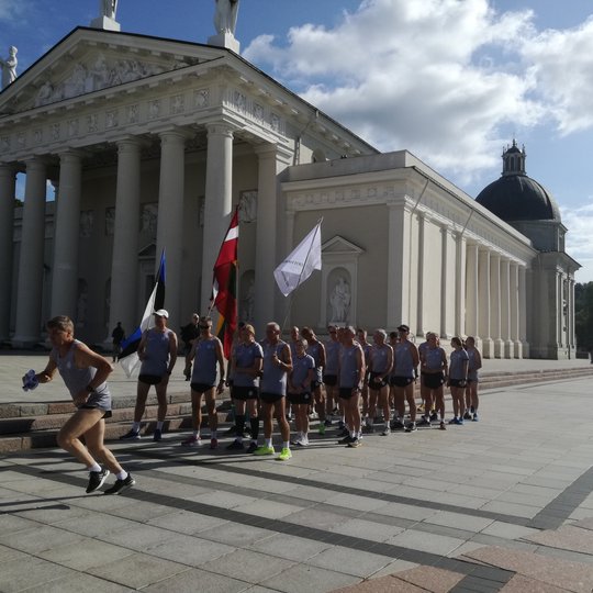 Tradiciško jubiliejinio estafetinio bėgimo dalyviai. Jie nuotraukoje pozuoja tarp Katedros varpinės ir Katedros. Daug vėliavų, vienoda bėgikų apranga, labai graži diena, daug saulės.