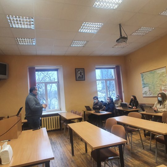 Paskaitų auditorija. Prie stalų susėdę studentai. Priešais juos stovi paskaitos lektorius. Ant sienų kabo keli žemėlapiai ir du televizoriai. Žmonės nuotraukoje dėvi veidą dengiančias medicinines kaukes. Yra dienos metas, tačiau auditorijoje įjungtas apšv