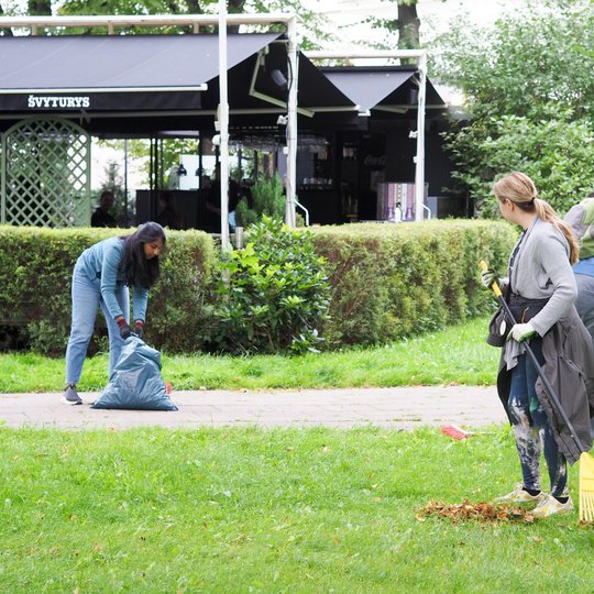 Žmonės parke tvarko aplinką – viena moteris grėbia lapus, kiti renka juos į maišą.