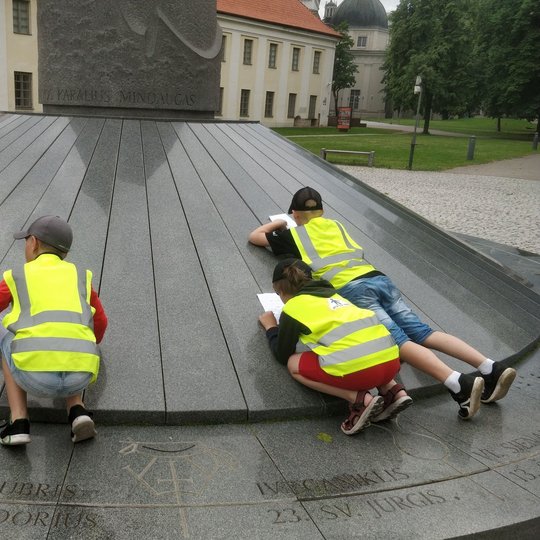 Vaikai apsirengę ryškiomis šviesą atspindinčiomis liemenėmis. Jie sprendžia orientacinio žaidimo užduotis. Vaikai sutūpę aplinkui Mindaugo paminklą.