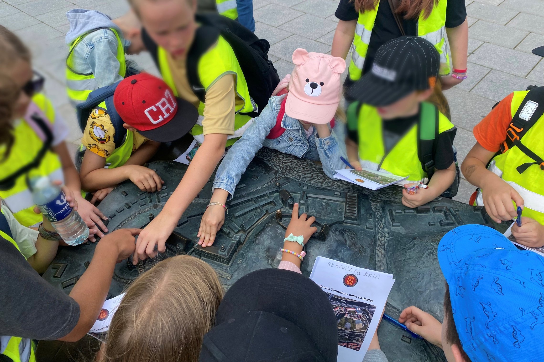Vaikai su ryškiomis liemenėmis tyrinėja Vilniaus Žemutinės pilies maketą lauke, dalyvaudami edukacijoje