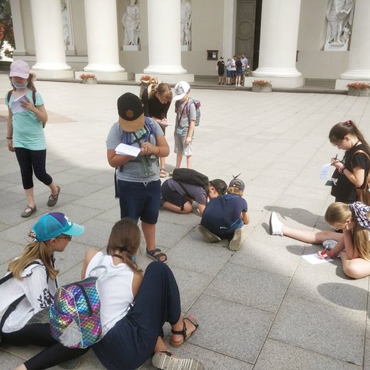 Būrelis vaikų su turistinėmis kuprinėmis priešais Vilniaus katedros fasadą sprendžia orientacinio žaidimo užduotis.