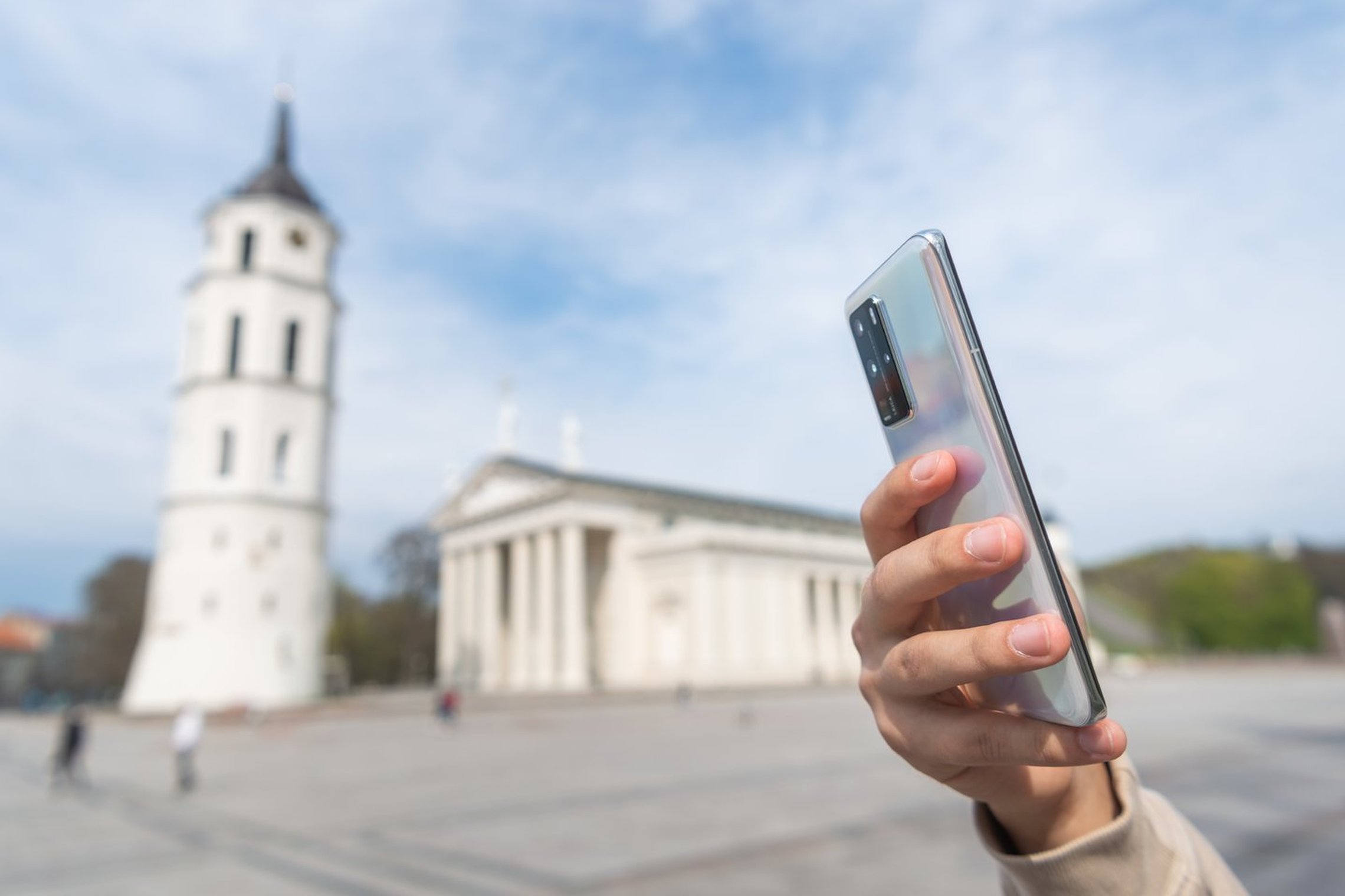 Nuotraukoje Viniaus katedra ir Katedros varpinė. Pirmame nuotraukos plane yra žmogaus ranka, kuri laiko mobilų telefoną.