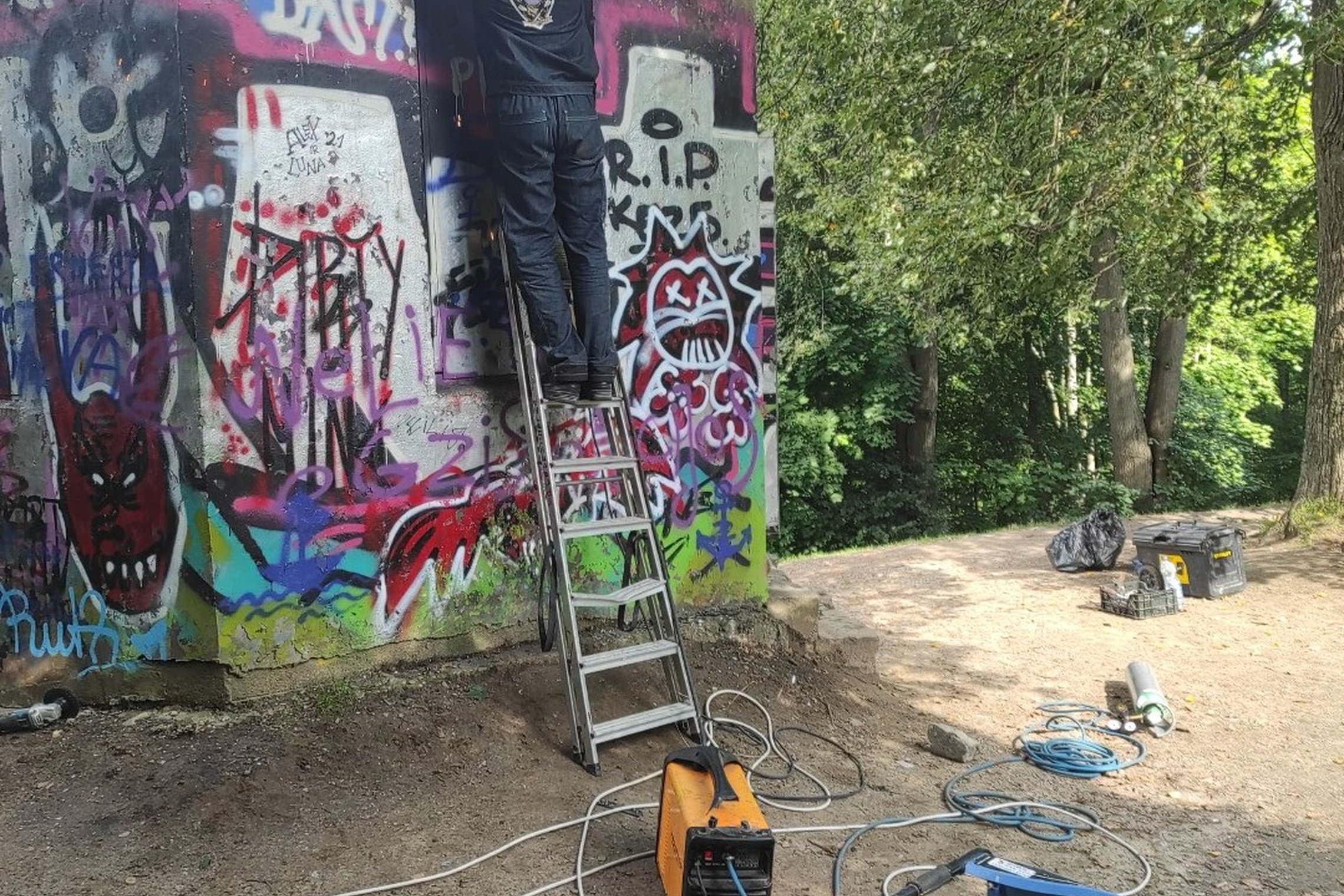 Darbininkas, pailipęs ant metalinių kopečių uždarinėja Altanos bokštelio duris. Ant žemės padėti darbo įrankiai. Pat bokštelis išpaišytas graffiti piešiniais.