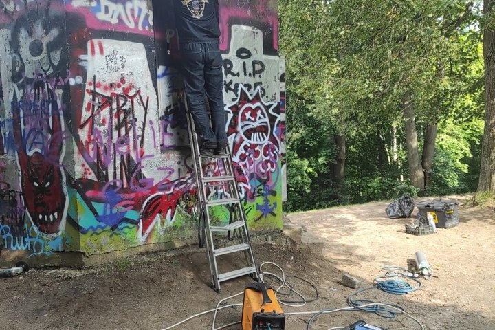 Darbininkas, pailipęs ant metalinių kopečių uždarinėja Altanos bokštelio duris. Ant žemės padėti darbo įrankiai. Pat bokštelis išpaišytas graffiti piešiniais.