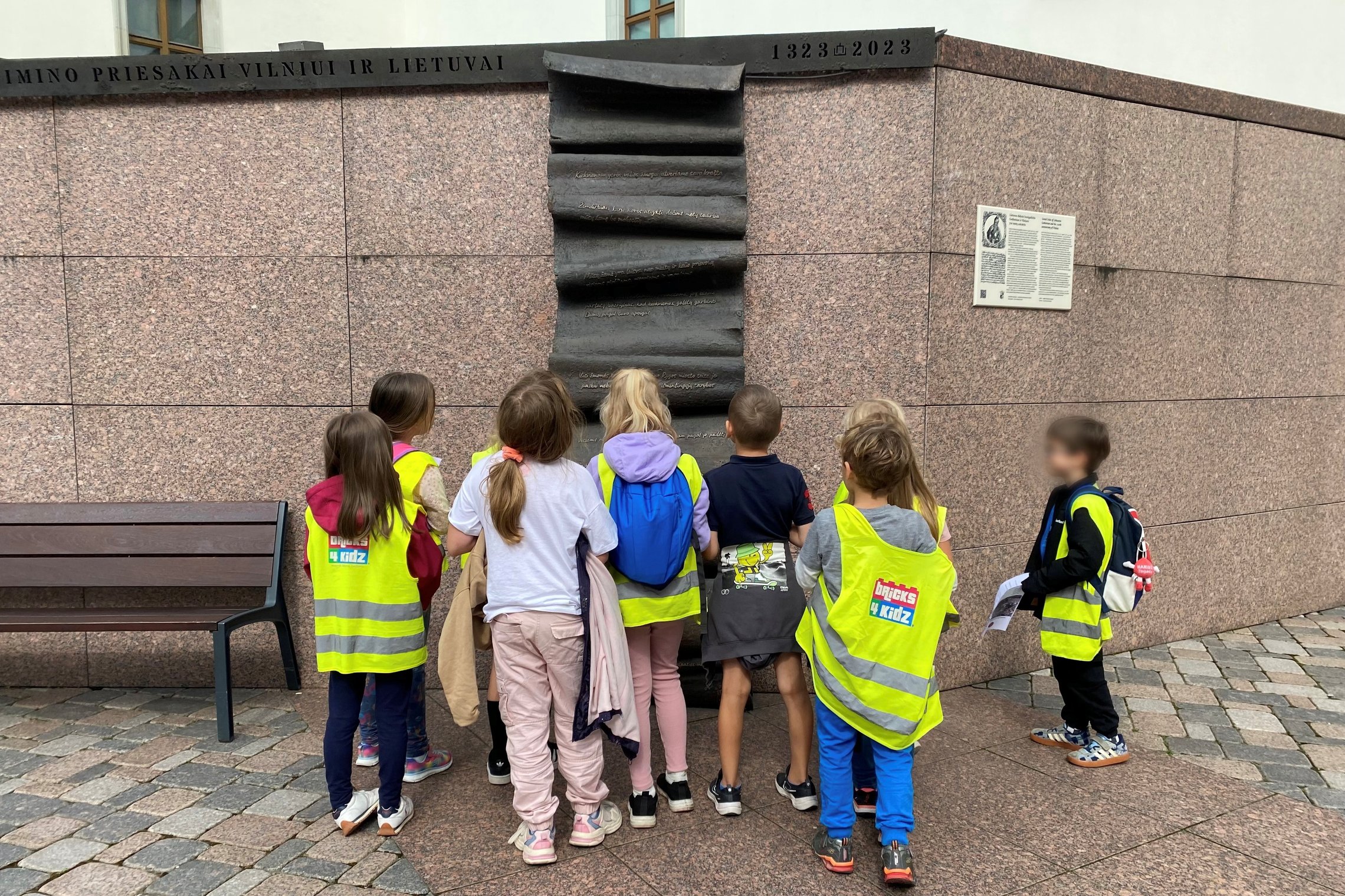 Bricks4kidz stovyklos vaikai, vilkintys ryškiaspalves liemenes, susidomėję žiūri į Gedimino laiškų paminklą Vilniuje