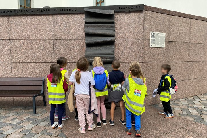 Bricks4kidz stovyklos vaikai, vilkintys ryškiaspalves liemenes, susidomėję žiūri į Gedimino laiškų paminklą Vilniuje