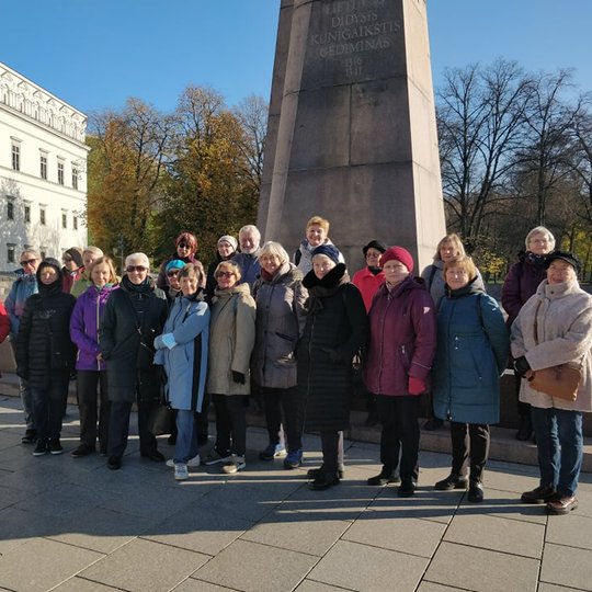 Ekskursijos dalyviai stovi prie Gedimino paminklo. Kairėje pusėje matosi Valdovų rūmų.