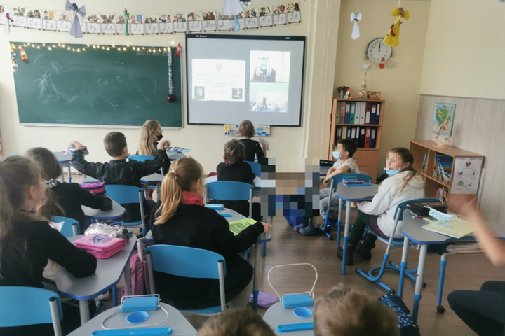 Pradinukų būrys stebi didelį ekraną. Vyksta nuotolinė edukacija. Kiekvienas vaikas sėdi prie jam skirto mokyklinio stalo. Šalai ekrano kabo klasės lenta.