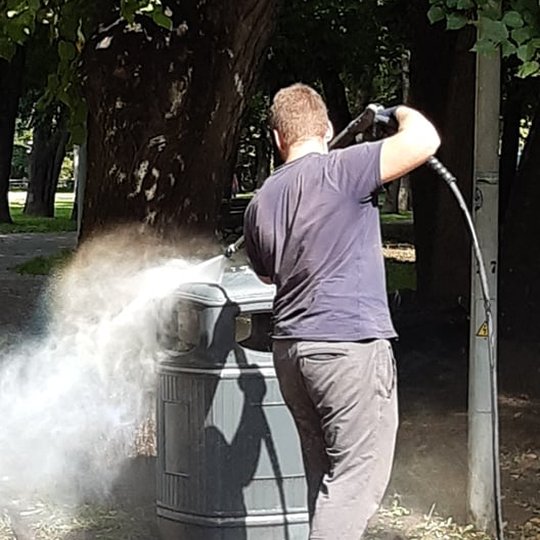 Jaunas vyriškis, apsirengęs tamsiai pilkais, trumpom rankovėm marškinėliais, tokios pat spalvos kelnėm, storapadžiais batais, su darbinėm pirštinėm laiko žarną su „pistoletu”. Didelio spaudimo vandens srovė nutaikyta į šiukšliadėžės paviršių, srovė tokia 
