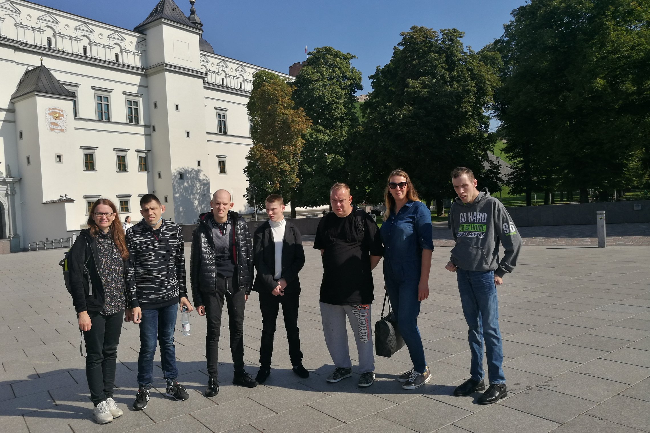 Kelių žmonių grupė stovi Vilniaus pilių rezervato teritorijoje. Už jų nugarų yra balti valdovų rūmai ir senas Pilių parkas.