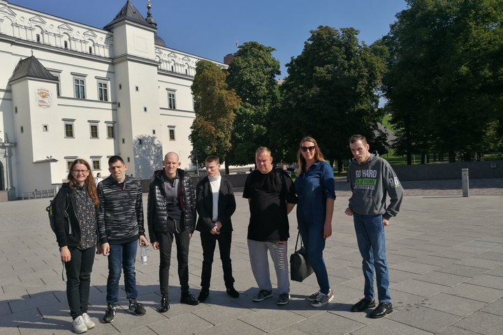 Kelių žmonių grupė stovi Vilniaus pilių rezervato teritorijoje. Už jų nugarų yra balti valdovų rūmai ir senas Pilių parkas.