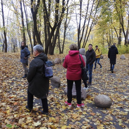 Ekskursijos dalyviai stovi senovės lietuvių tikėjimo apeigų šventvietėje. Prikritę daug lapų, graži rudens diena. Aplink ritualų ratą išdėlioti akmenys.