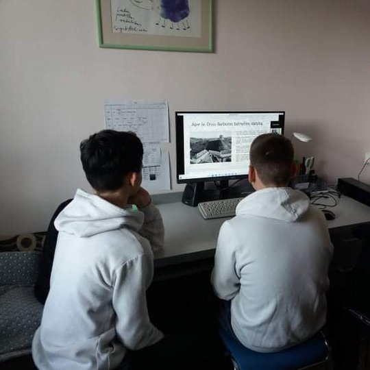 Trys vaikai prie stalo žiūri į televizoriaus ekraną. Vyksta nuotolinė edukacija. Vaikai dėvi bliuzonus su kapišonais.