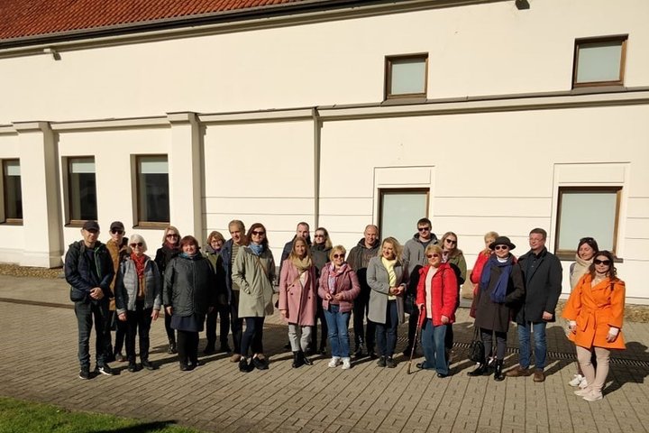 Grupė žmonių stovi prie namo sienos, daugelis yra su tamsiais akiniais. Žmonės atsisegę savo striukes ir paltus. Yra graži saulėta diena.