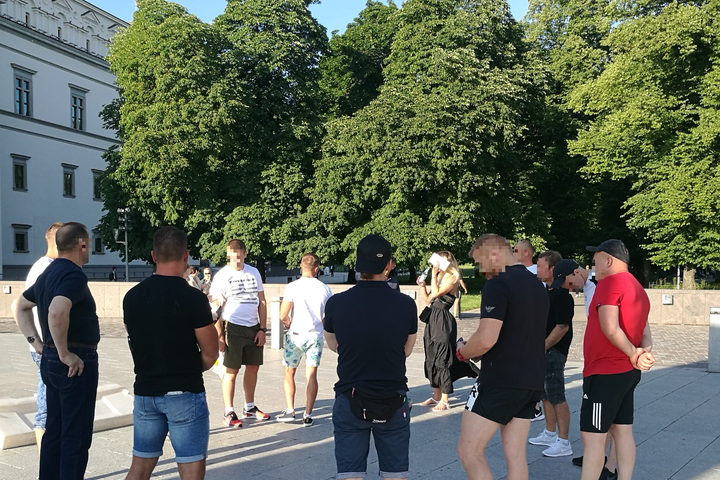 Grupė ekskursijos dalyviai stovi ratu Valdovų rūmų fone. Nuotraukos centre matosi Pilių parko medžiai.