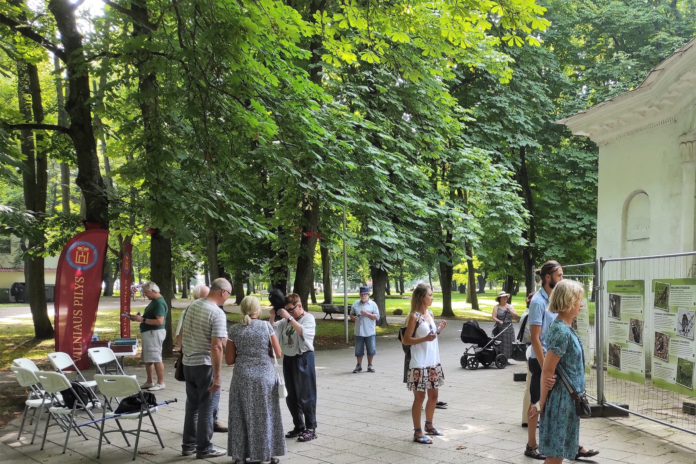 Pilių parke vyksta Vilniaus pilių paukščių nuotraukų parodos atidarymas. Susirinkę būrelis žmonių. Yra išdėliotos kėdės ir stalas. Aplinkui seni medžiai.