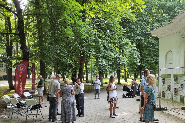 Pilių parke vyksta Vilniaus pilių paukščių nuotraukų parodos atidarymas. Susirinkę būrelis žmonių. Yra išdėliotos kėdės ir stalas. Aplinkui seni medžiai.