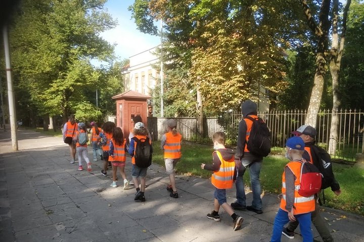 Palei gatvę eina būrelis vaikų. Jie eina poromis, išsirikiavę vienas už kito. Vyresni vaikai veda mažesnius. Visi dėvi šviesą atspindinčias apsauginęs liemenes. Graži vasaros diena, šviečia saulė.