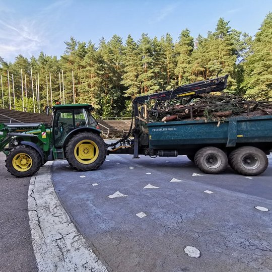 Didžiulis traktorius su priekaba, kurioje yra nupjauta mediena, kurią ruošiasi išvežti. Traktorius stovi Dainų slėnio slėnio estradoje.
