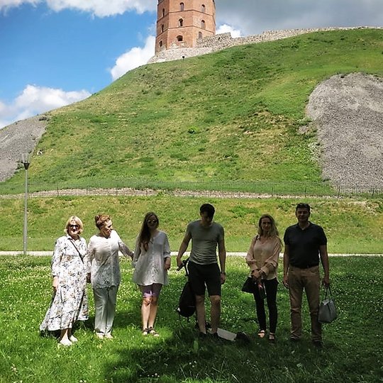 Šešių žmonių grupelė Gedimino kalno papėdėje. Virš jų stūkso Gedimino piies gynybinis bokštas. Žmonės su vasariniais rūbais, yra šilta.