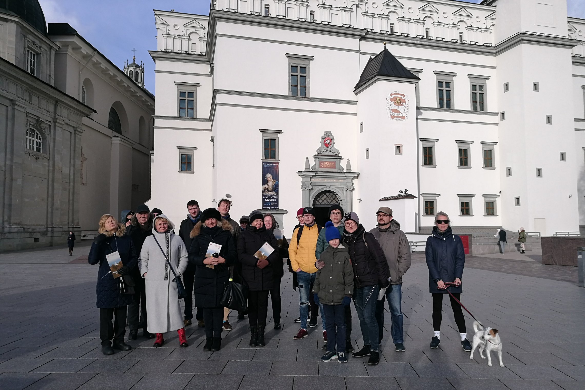 Ekskursijos didelė žmonių grupė pozuoja nuotraukai Valdovų rūmų fone. Tarp ekskursijos dalyvių yra ir šuo. Saulėta žiemos diena, vaiskus dangus.