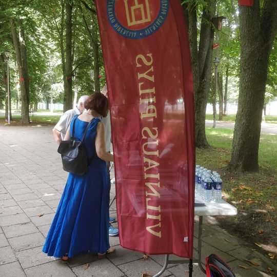 Pilių parke vyksta Vilniaus pilių paukščių nuotraukų parodos atidarymas. Susirinkę būrelis žmonių. Yra išdėliotos kėdės ir stalas. Aplinkui seni medžiai. Pirmame nuotraukos plane plevesuoja Vilniaus pilių rezervato vėliavos.