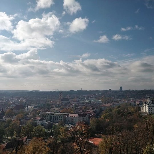Nuostabi ir šilta rudens diena ir nuotraukoje daug vaiskaus dangaus. Keli balti debesėliai. Vaizdas nuo Trijų kryžių kalno. Matosi Gedimino pilis, Valdovų rūmai ir visas miestas. Nuotraukos pirmame plane - daug medžių. Jie dar pasidabinę rudens spavomis.
