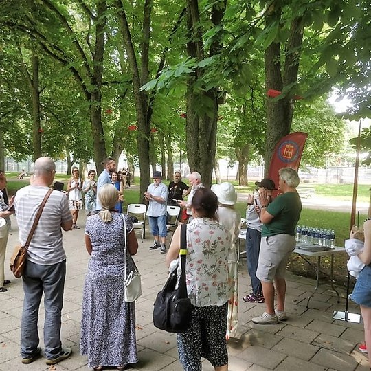 Pilių parke vyksta Vilniaus pilių paukščių nuotraukų parodos atidarymas. Susirinkę būrelis žmonių. Yra išdėliotos kėdės ir stalas. Aplinkui seni medžiai.