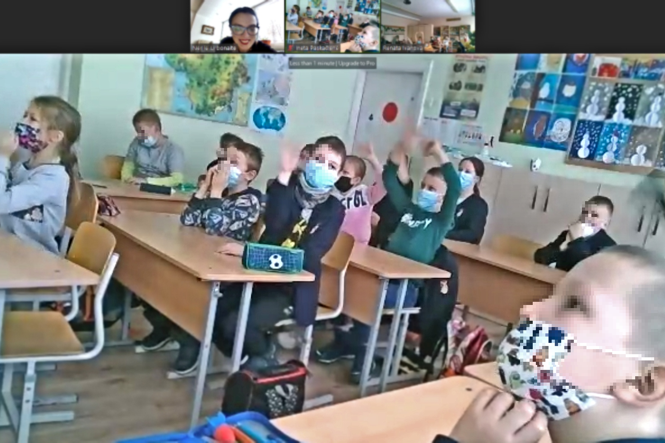Vyksta nuotolinė edukacija, nuotraukoje daug vaikų, kurie sėdi suoluose. Beveik visi su medicinėmis veido kaukėmis. Keli vaikai mojuoja rankomis. Ant sienų dau vaikų piešinių, kabo ir žemėlapis. Virtualiame susitikime dalyvauja dvi klasės.