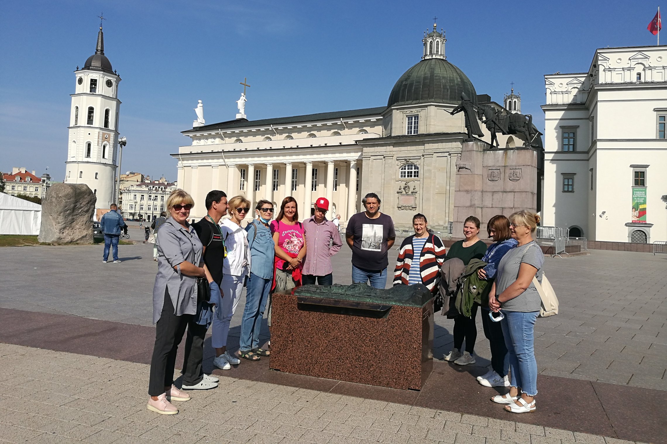 Grupė ekskursijos dalyvių stovi aplink Vilniaus pilių maketą Katedros aikštėje, netoli Gedimino paminklo. Žmonių fone Vilniaus katerda, varpinė ir Valdovų rūmų kampas.