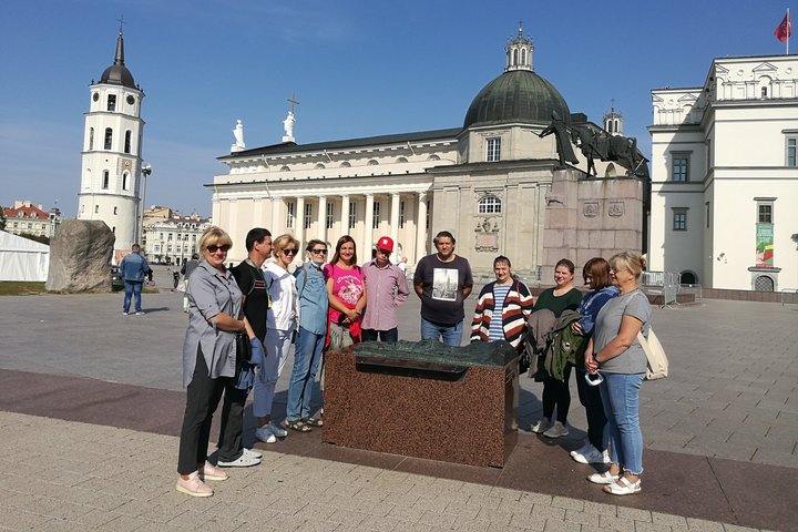Grupė ekskursijos dalyvių stovi aplink Vilniaus pilių maketą Katedros aikštėje, netoli Gedimino paminklo. Žmonių fone Vilniaus katerda, varpinė ir Valdovų rūmų kampas.